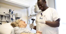 A male nurse in white scrubs smiles while examining an older patient who is lying in a hospital bed and looking up at him