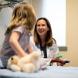 A female nurse practitioner in a white coat smiles while talking with a young female child sitting on an exam table, who is holding a stuffed animal.