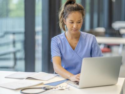nurse typing on laptop with papers on desk in front of her