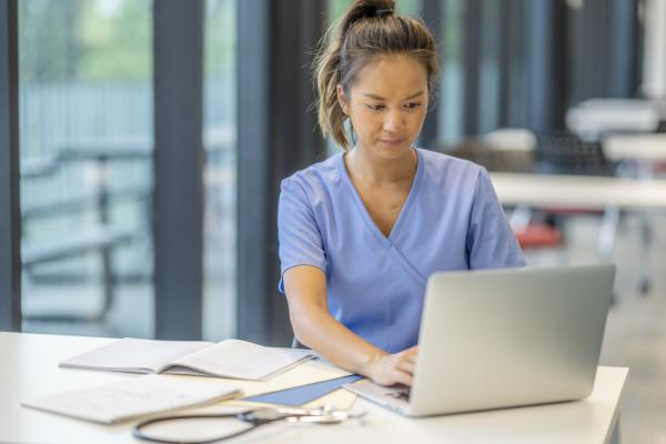 nurse typing on laptop with papers on desk in front of her